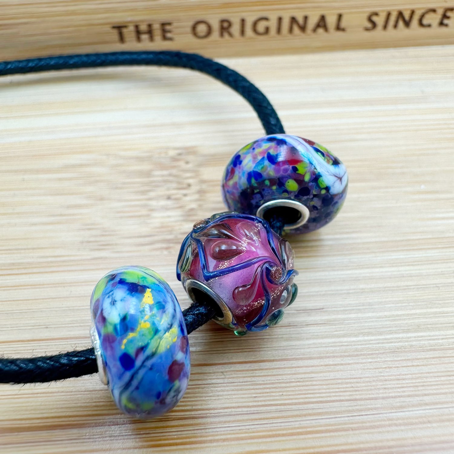 Bracciale Alchimia composto da: 2 beads unici multicolor laterali e un beads unico centrale con palette di colori: rosa ametista, prugna, blu zaffiro e riflessi di bronzo. | Golden Party Trollbeads